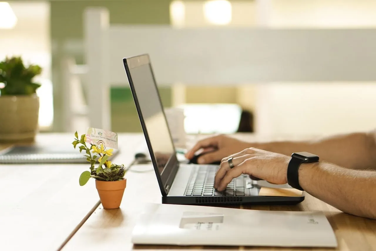 Mãos digitando em um notebook sobre uma mesa com plantas